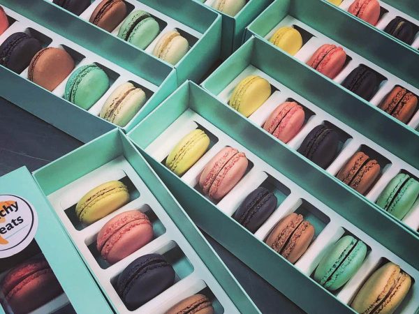 Boxes of colorful macarons in various flavors are neatly arranged in rows. Each box has a transparent lid and compartments for individual macarons. A label on the box reads Frenchy Treats.