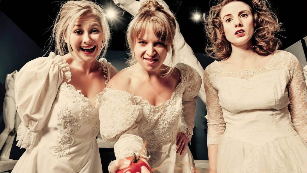 looking at the camera. The middle woman holds a red apple. The background is dimly lit with abstract white shapes hanging above them.