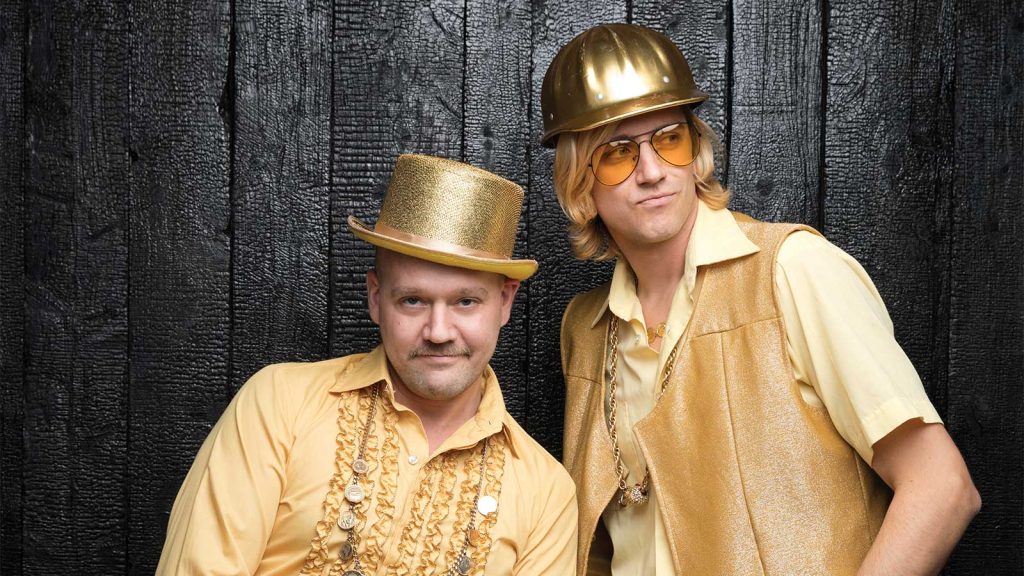 the other in a shiny hard hat and vest. Both have playful expressions and layered gold chains.