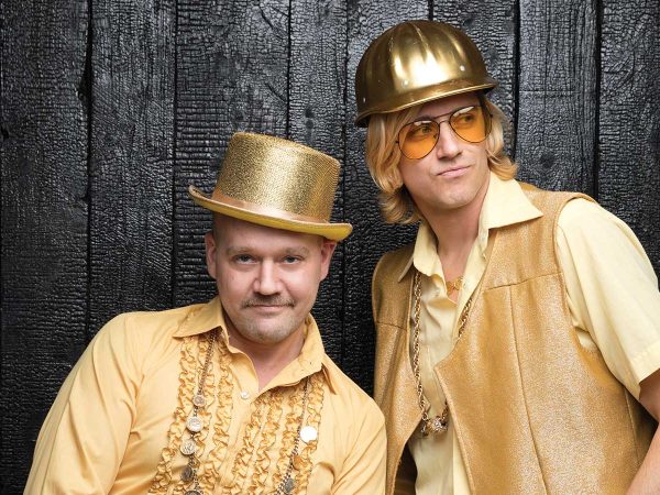 the other in a shiny hard hat and vest. Both have playful expressions and layered gold chains.