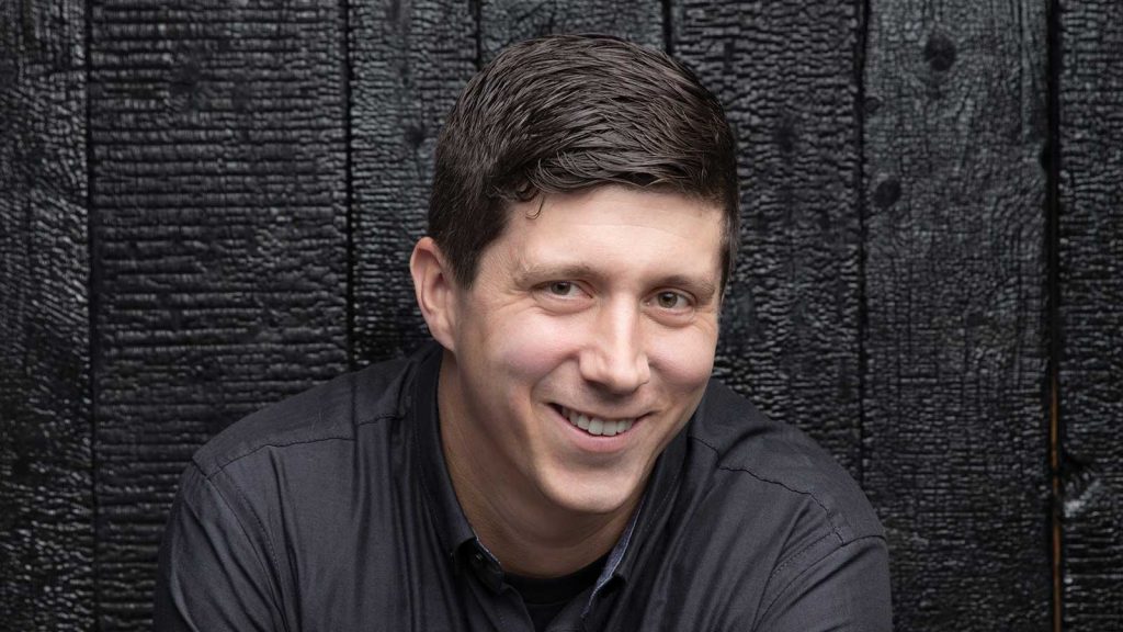 dark hair wearing a dark shirt against a textured dark wood background.
