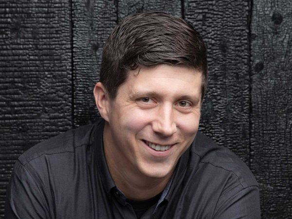 dark hair wearing a dark shirt against a textured dark wood background.