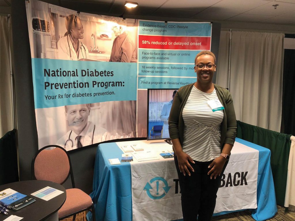 A woman stands smiling in front of a booth for the National Diabetes Prevention Program. The booth includes a banner promoting diabetes prevention and a table with informational materials. The setting appears to be a conference or event.