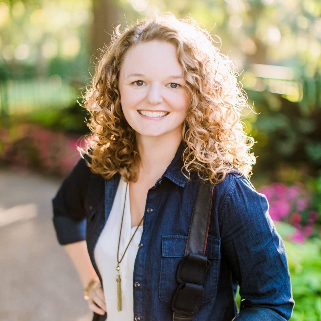 A person with curly hair smiles while standing outdoors in a sunlit park. Theyre wearing a denim jacket over a white shirt with a pendant necklace. The background features green trees and pink flowers.