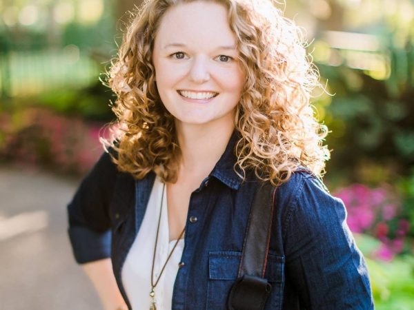 A person with curly hair smiles while standing outdoors in a sunlit park. Theyre wearing a denim jacket over a white shirt with a pendant necklace. The background features green trees and pink flowers.