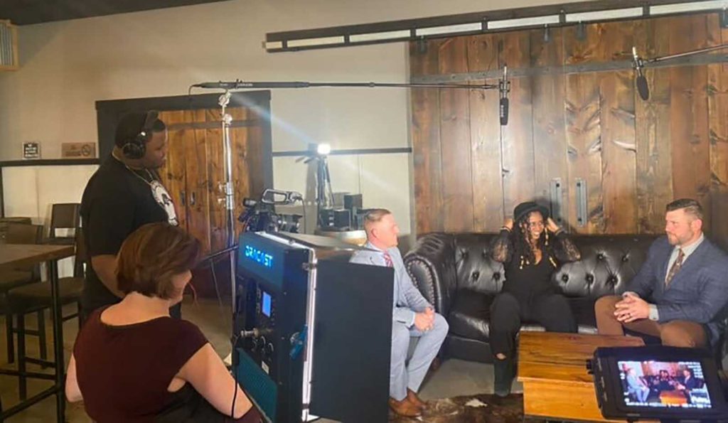 Two people filming and interviewing three people sitting on a couch with large wooden sliding barn doors behind them