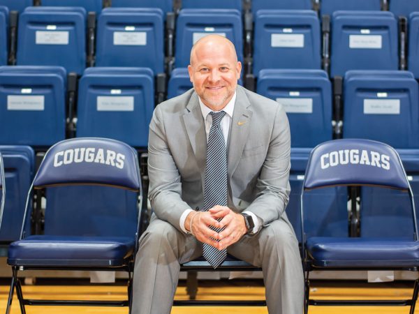 smiling at the camera. The seat is part of a row of chairs labeled COUGARS. Behind him are rows of blue stadium seats.