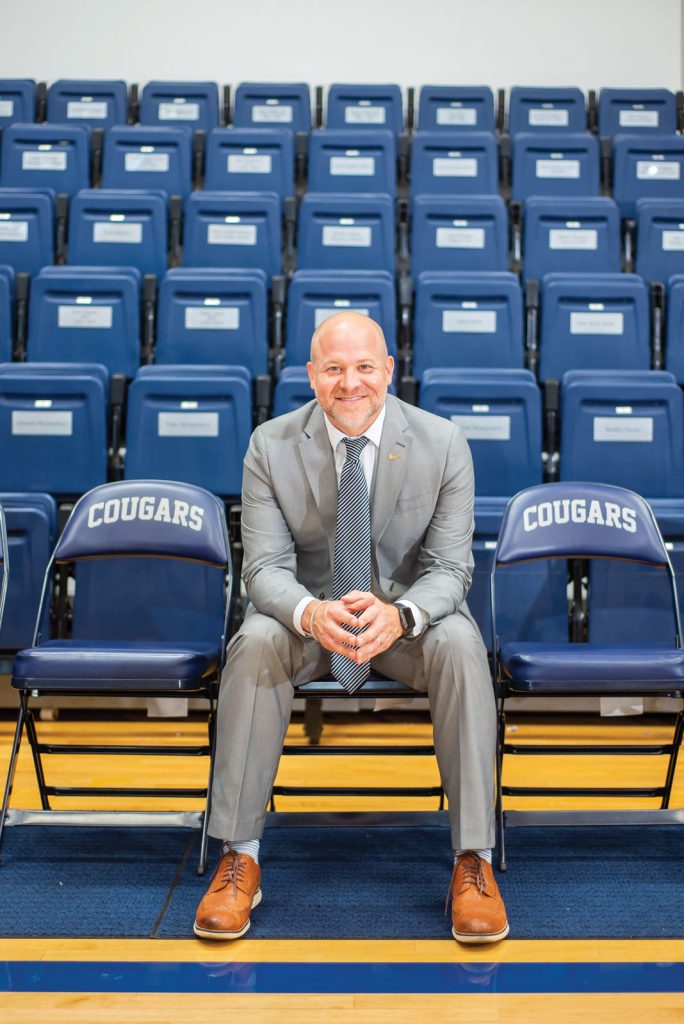smiling at the camera. The seat is part of a row of chairs labeled COUGARS. Behind him are rows of blue stadium seats.