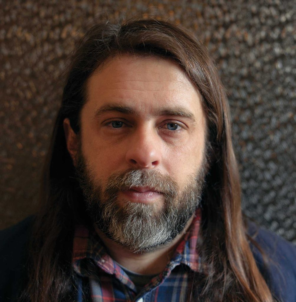 wearing a plaid shirt and dark jacket. He is facing the camera with a neutral expression. The background is a textured