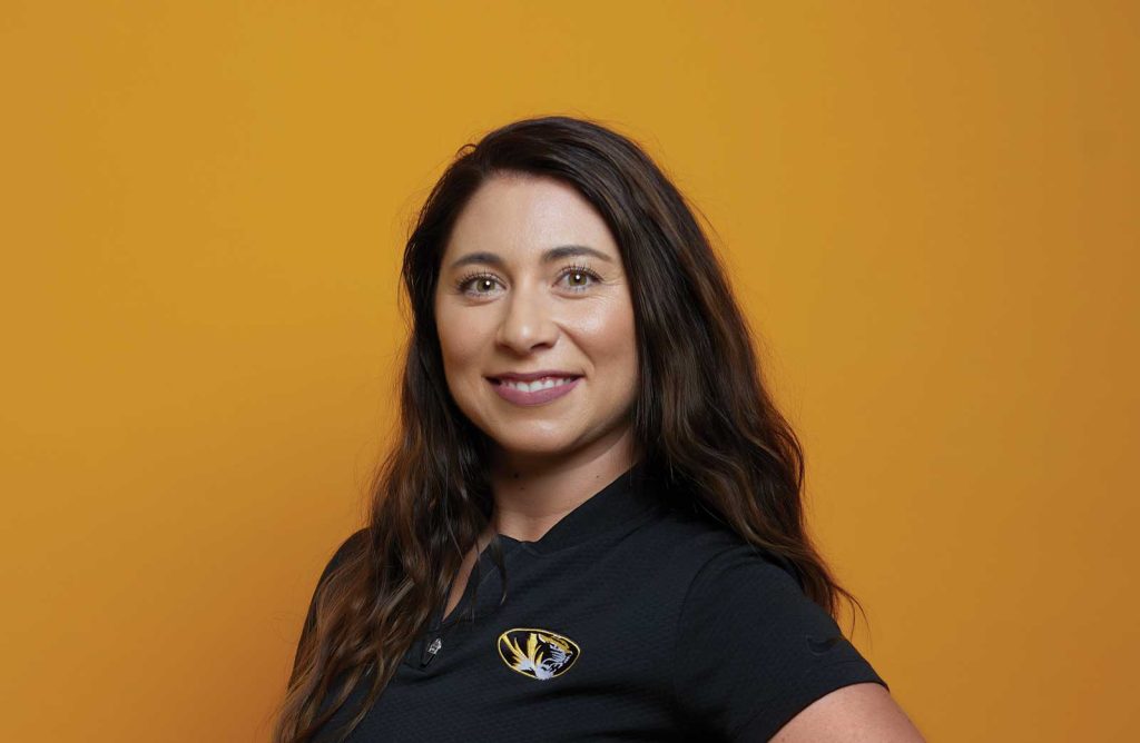 A person with long brown hair smiles warmly against an orange background. They are wearing a black shirt with a tiger logo.