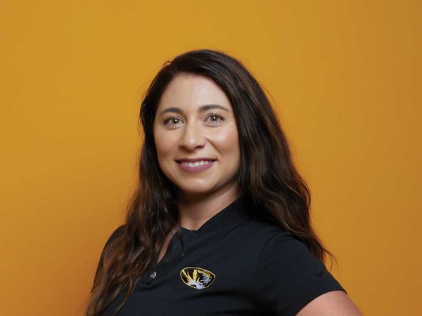 A person with long brown hair smiles warmly against an orange background. They are wearing a black shirt with a tiger logo.