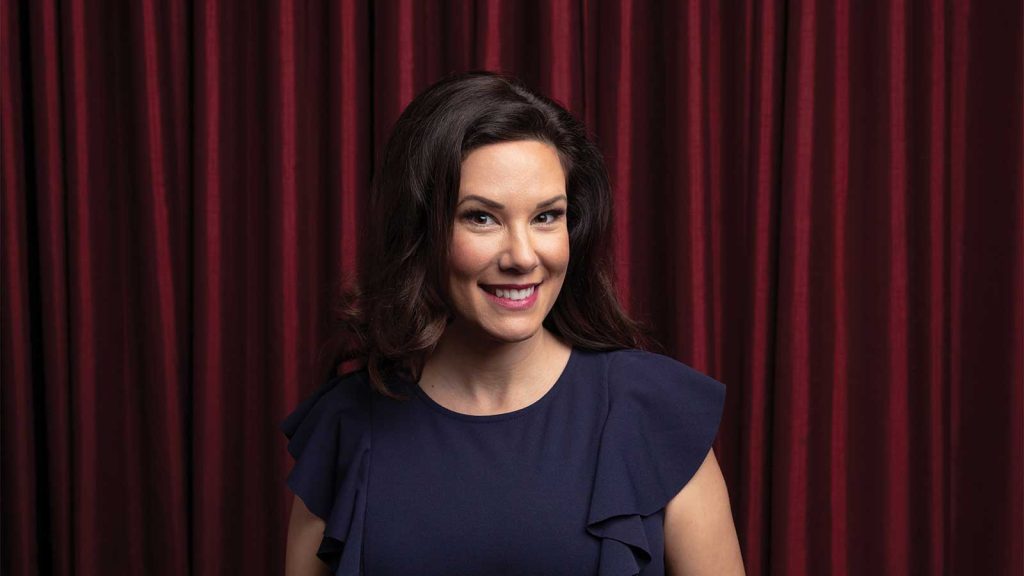 A person with long dark hair and a blue dress stands smiling in front of deep red curtains.