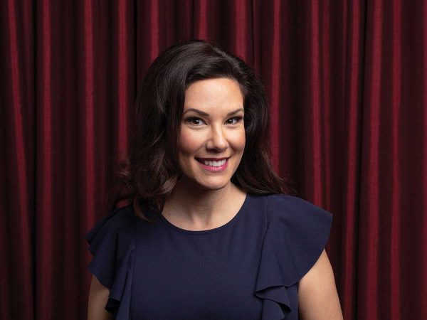 A person with long dark hair and a blue dress stands smiling in front of deep red curtains.