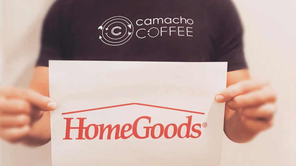 A person in a black camacho COFFEE t-shirt holds a paper with the HomeGoods logo. The background is plain and blurry.