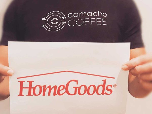 A person in a black camacho COFFEE t-shirt holds a paper with the HomeGoods logo. The background is plain and blurry.