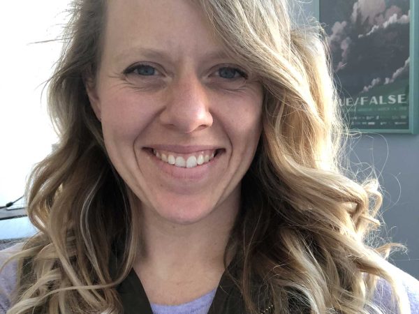 curly blonde hair wearing a purple shirt and dark vest. They are indoors with a poster on the wall in the background.
