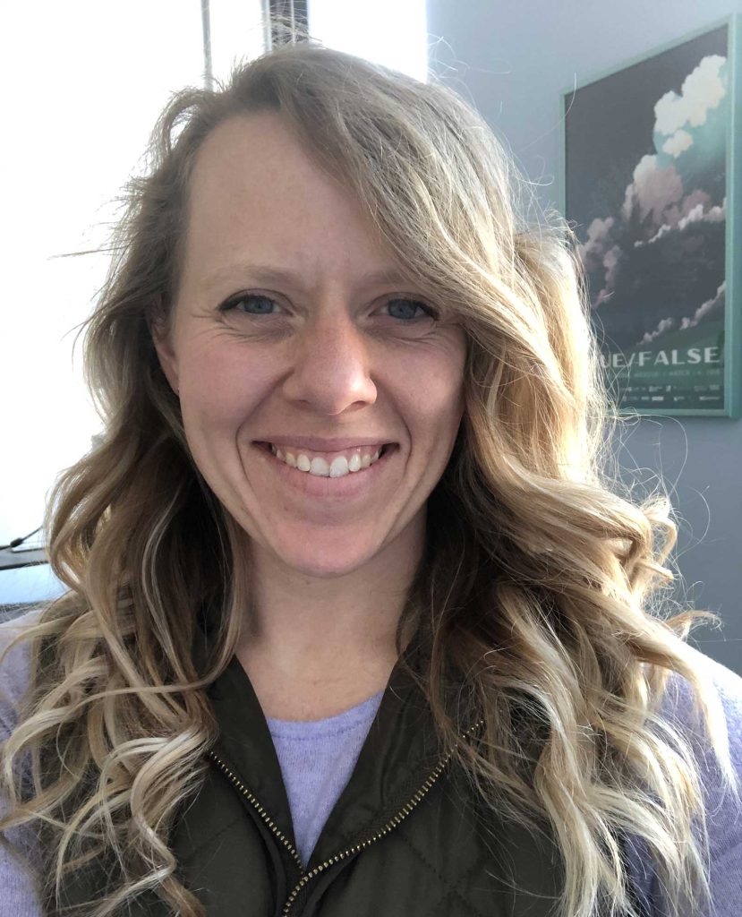curly blonde hair wearing a purple shirt and dark vest. They are indoors with a poster on the wall in the background.