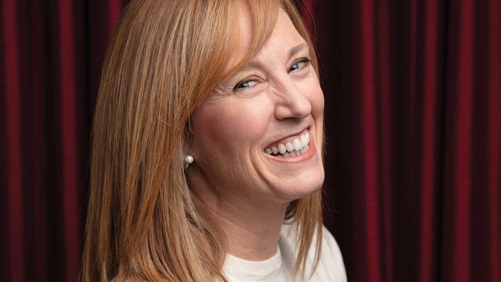 light brown hair and wearing a white top looks over her shoulder against a rich burgundy curtain backdrop. She is wearing pearl earrings and appears joyful.