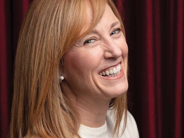 light brown hair and wearing a white top looks over her shoulder against a rich burgundy curtain backdrop. She is wearing pearl earrings and appears joyful.