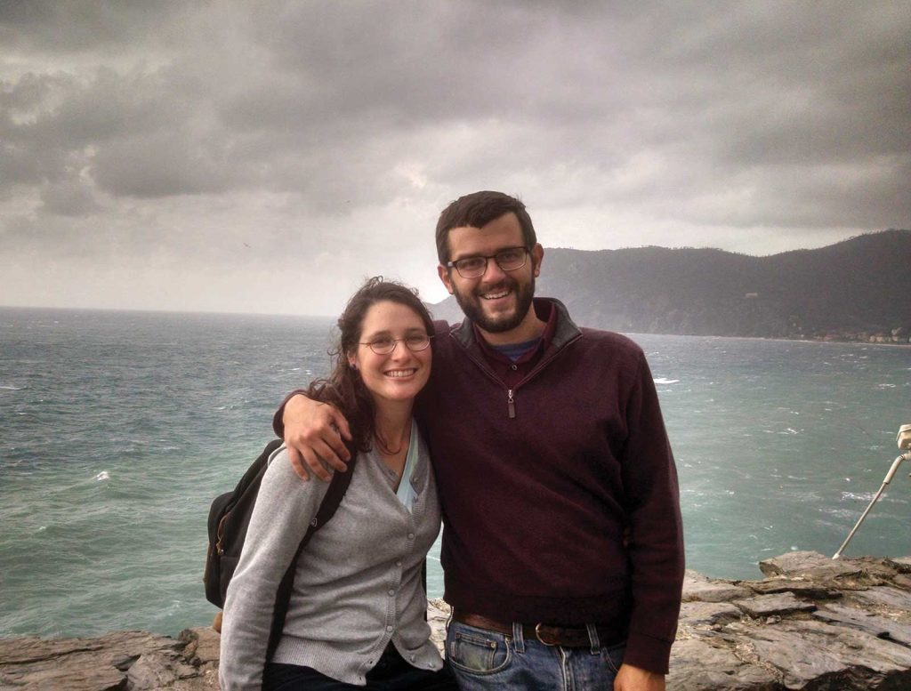 A smiling couple stands on a rocky coastline with an overcast sky and ocean in the background. The man has his arm around the womans shoulder. Both are wearing casual clothing and glasses.
