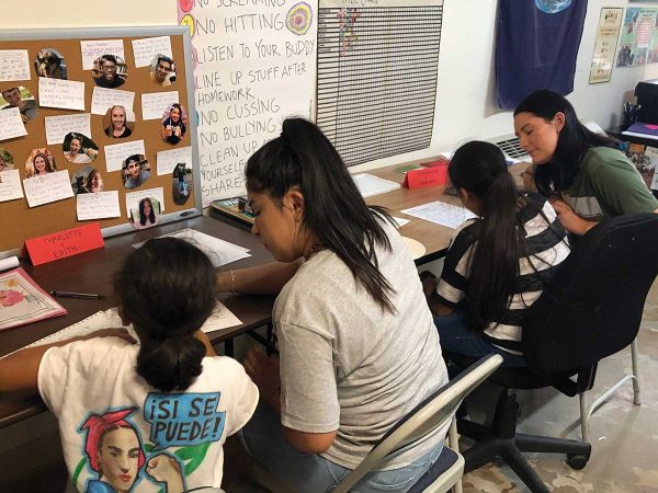 accompanied by an adult who is assisting them. Notes and photos are pinned to a bulletin board on the wall. Snacks are on the table. The room has a casual