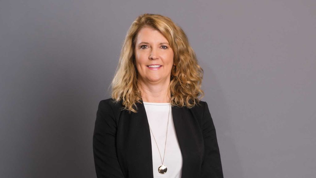curly blonde hair smiling at the camera. She is wearing a black blazer over a white top and a long necklace with a pendant. The background is plain grey.