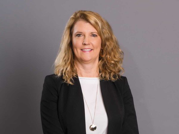 curly blonde hair smiling at the camera. She is wearing a black blazer over a white top and a long necklace with a pendant. The background is plain grey.