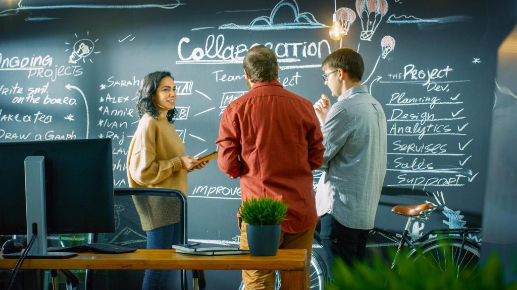Three people engage in discussion in front of a blackboard filled with writing and drawings about collaboration and project management. A computer and potted plants are on a table in the foreground.