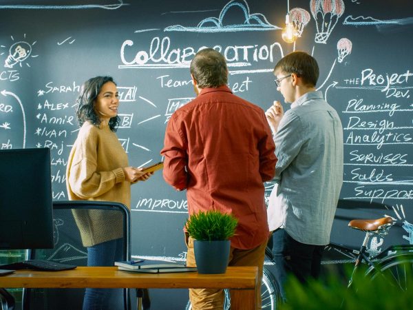 Three people engage in discussion in front of a blackboard filled with writing and drawings about collaboration and project management. A computer and potted plants are on a table in the foreground.