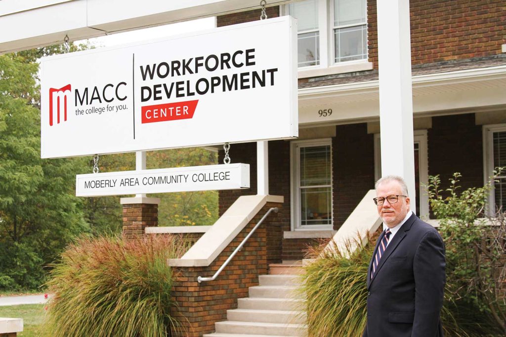 posing beside a large sign displaying the college's name.