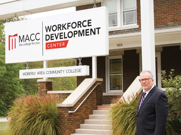 posing beside a large sign displaying the college's name.