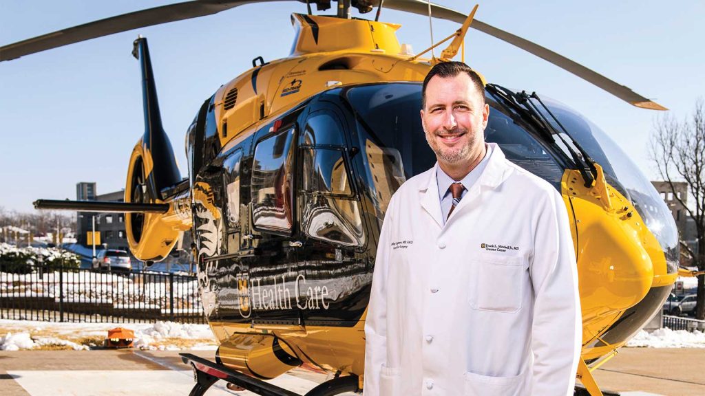 A man in a white medical coat stands smiling in front of a yellow and black helicopter. The background features a clear sky and a snowy ground.