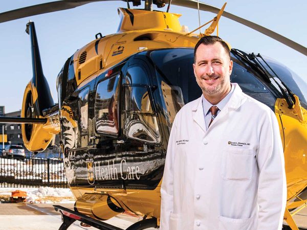 A man in a white medical coat stands smiling in front of a yellow and black helicopter. The background features a clear sky and a snowy ground.