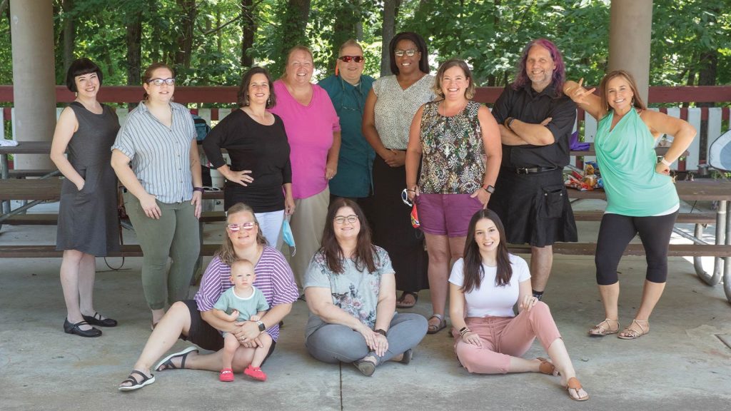 while others stand behind them. Trees and picnic tables are visible in the background. One person holds a baby.