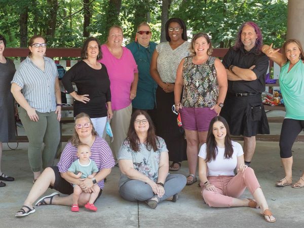 while others stand behind them. Trees and picnic tables are visible in the background. One person holds a baby.