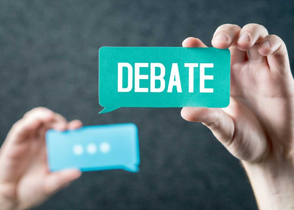one hand holding up a text bubble shaped card with the text "Debate" and a hand holding up a text bubble card with the text "..." in the background