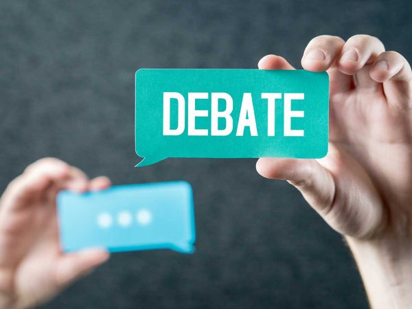 one hand holding up a text bubble shaped card with the text "Debate" and a hand holding up a text bubble card with the text "..." in the background