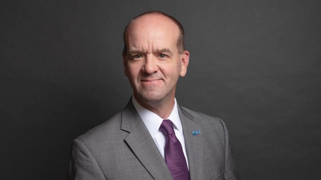 A man in a gray suit with a purple tie stands against a dark gray background. He is smiling slightly and wearing a lapel pin on his suit jacket.