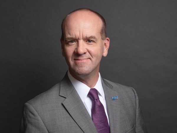 A man in a gray suit with a purple tie stands against a dark gray background. He is smiling slightly and wearing a lapel pin on his suit jacket.