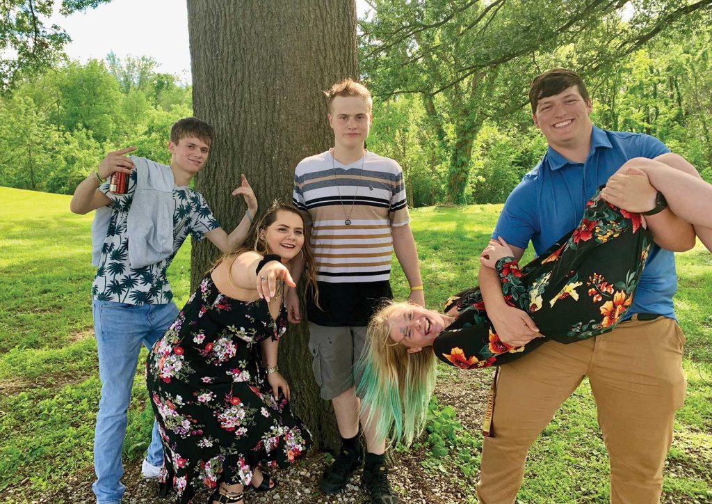 and another is playfully lifting a person wearing a floral dress. The background is a grassy area with trees under a sunny sky. Everyone appears to be enjoying themselves.