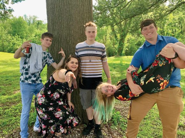 and another is playfully lifting a person wearing a floral dress. The background is a grassy area with trees under a sunny sky. Everyone appears to be enjoying themselves.