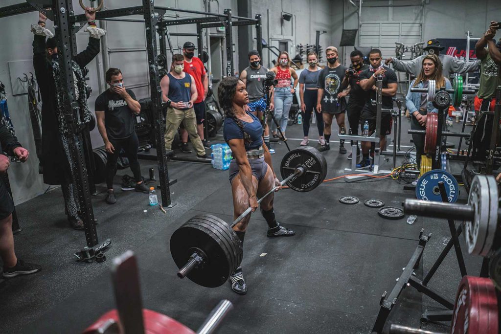 surrounded by an audience of onlookers. Some spectators hold smartphones to capture the moment. Weightlifting equipment and gym accessories are visible in the background.