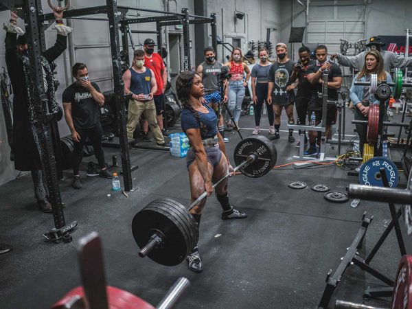 surrounded by an audience of onlookers. Some spectators hold smartphones to capture the moment. Weightlifting equipment and gym accessories are visible in the background.