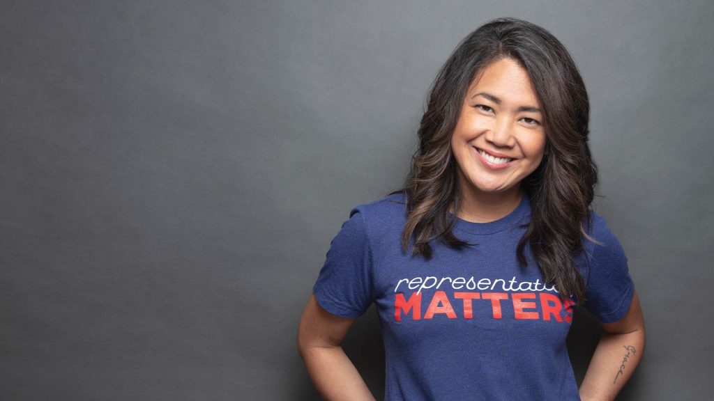 wavy hair smiles in front of a gray background. They are wearing a blue t-shirt with the phrase representation MATTERS in bold red and white letters.