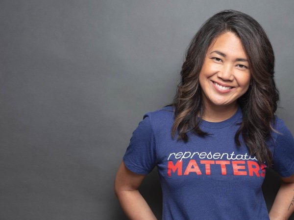 wavy hair smiles in front of a gray background. They are wearing a blue t-shirt with the phrase representation MATTERS in bold red and white letters.