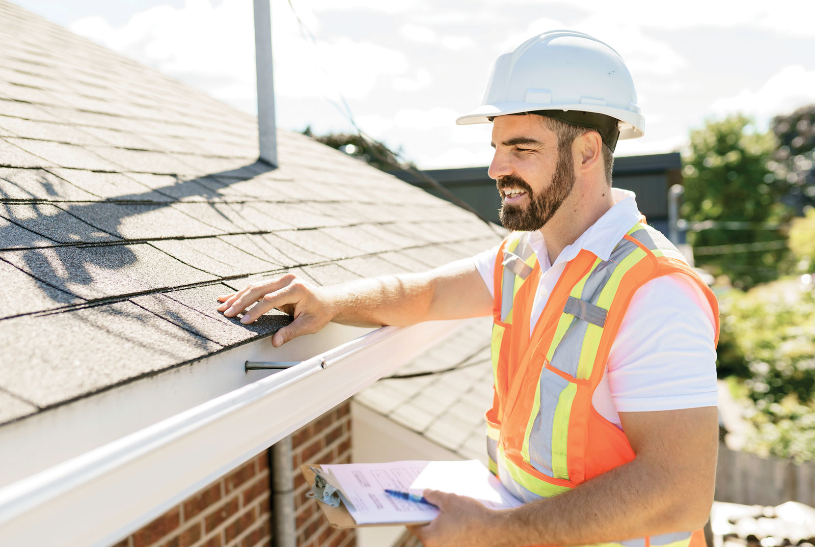 Man in a hardhat and orange vest inspecting a roof.