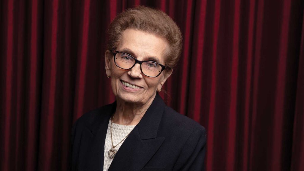 An older woman with short brown hair and glasses smiles while wearing a black blazer over a white top. She stands in front of a red curtain.