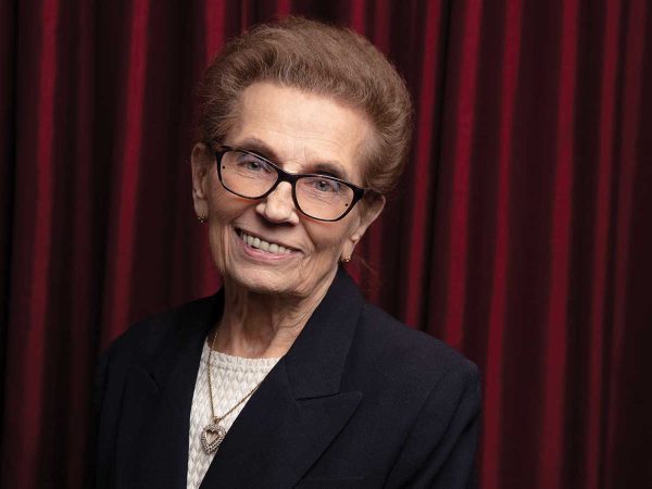 An older woman with short brown hair and glasses smiles while wearing a black blazer over a white top. She stands in front of a red curtain.
