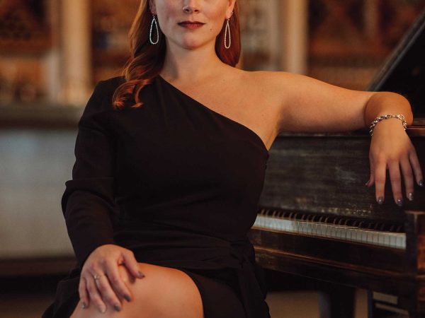 Headshot of a women named Michele Curry sitting on a piano bench and resting her arm on a black piano