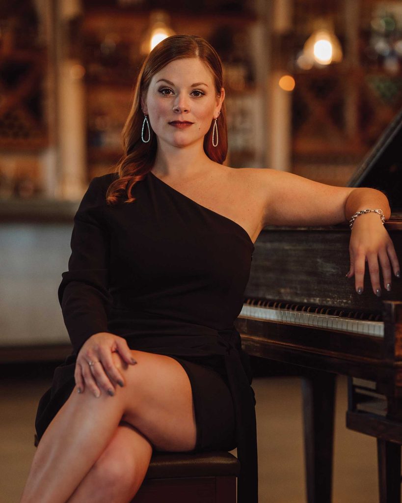 Headshot of a women named Michele Curry sitting on a piano bench and resting her arm on a black piano